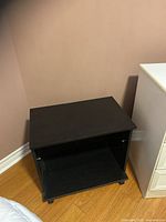 Black rolling side table, rectangular shape, situated on wooden floor against beige wall. Shows open shelf with black backing.