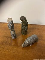 Photo showing three stone carvings: two human figure carvings and one rhino figure with chipped horn on a wooden surface.