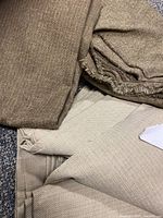Image showing a light brown textured upholstery fabric rolled or folded and a lighter beige fabric underneath, both lying on a carpet.