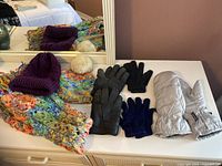 Overview showing purple knit Auclair hat with pompom detached, colorful knit scarf and matching knit hat, sheepskin gloves, thin gloves and Thinsulate mittens arranged on white surface.