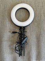 Photo showing USB powered ring light with white diffuser, attached black USB cable, remote control, and tripod stand placed on textured surface.