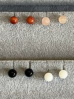 Photo showing six pairs of small round stone stud earrings in orange, pale pink, black, and white, laid out in two rows on a textured surface.