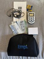 Assortment of items including the white handheld Empi muscle stimulator device with control buttons and screen, electrode wires, multiple adhesive electrode pad packages, batteries, user manual, small white box, and black soft carrying case with Empi logo, all laid out on gray fabric surface.