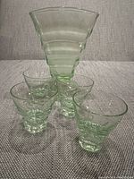 Green fan vase and four green stemless glasses arranged in front of a neutral backdrop on a soft surface