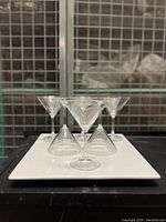 Six clear martini glasses arranged on a white square ceramic tray with a metal wire background, showing overall set layout.
