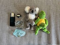 Photo showing all items in the lot arranged on fabric: Bulgari cologne, sea turtle plush, koala plush, glass card holder, glass candle holder, sea turtle carving