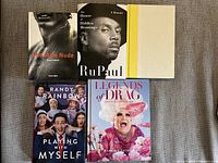 Top down view of five hardcover books arranged on a surface showing front covers. Titles include RuPaul memoir and queer themed books.