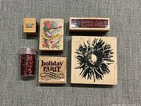 Top angle view of the front side of six wood backed rubber stamps and small container of red glitter, showing various stamp designs including text, floral, bird, and cake slice.