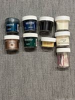 Top view of 9 small jars of embossing powders in various colors arranged in rows on grey fabric surface.