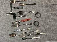 Photo of various silver plate cutlery, including forks, spoons, and smaller forks with white handles, alongside two napkin rings and an MCM red-handled can opener.