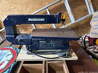 Photo of used blue Mastercraft scroll saw with blade attached and power cord wrapped below. Mounted on a wooden board.