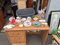 Overview of assorted china and figurines including plates, cups, teapot, and figurines on wooden desk.
