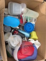 Photo of various plastic storage containers and lids, measuring cups and other kitchen utensils in a cardboard box.