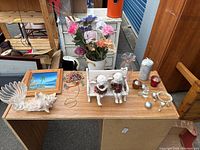 Top-down view of assorted decorative pieces on a wooden table including artificial flowers, ceramic sheep figurines, framed beach painting, glass vase, small decor items.