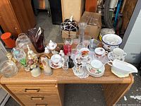 Top-down view of assorted glass and ceramic items arranged on a wooden desk showing bowls, vases, figurines, plates, and stemware.