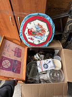 Various houseware items including a Santa Claus themed wall clock with a padded fabric border, glass bowls with lids, ceramic mugs, and some boxed items, some in plastic storage containers photographed from above.