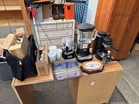 View of various kitchenware on wooden boards including two electric blenders, four white ceramic mugs, two espresso makers, decorative ceramic plate, wooden plate, and black carry bag with boxes.