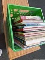Top-down and angled views showing the assortment of 12 books inside a green plastic crate, titles, colors, and book condition visible.