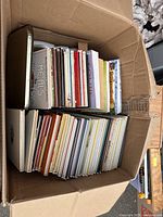 Box filled with cookbooks and recipe books viewed from above showing a variety arranged vertically and flat down in a cardboard box.