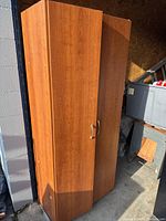 Photo of closed wood-finish cabinet with two doors, silver handles, standing upright in a garage or storage area.
