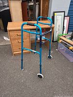 Blue foldable walker standing on pavement with wooden furniture and framed pictures in the background.
