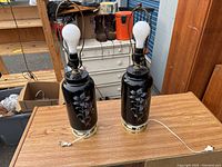 Pair of black ceramic lamps without shades, placed on wooden table. Lamps have floral design and attached white power cords with plugs.