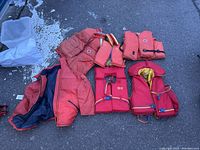 Photo showing six red lifejackets on the ground with various styles and inner linings visible.