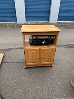 Front view of the light wood microwave stand with cabinet. Three VHS/DVD players stacked in open shelf beneath top surface.