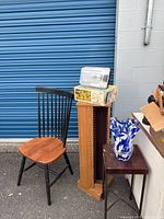 Wooden chair, tall media rack, small side table, blue and white glass vase, and unopened floodlight box against blue storage door backdrop