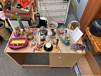 Top view of a wooden table covered with assorted candles, decorative bowls, glassware, and small household items, with a white dresser and other furniture in the background.