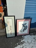 Two framed pictures leaning against a wall outside, one vertical with animal prints and one square styled bird print with pink mat