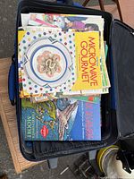 Open medium soft-sided suitcase filled with assorted cookbooks stacked inside, showing the cover of Microwave Gourmet on top.