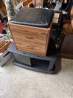 Photo showing wooden footstool with two drawers and cushioned top placed on top of a black TV stand with glass doors and open storage compartments.