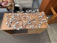 Top-down view of the metal wall art sculpture laid flat on a wooden desk. The sculpture depicts two trees with circular mirrors on branches.