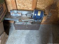Woodworking tool on metal stand with blue motor attached, rusted metal carry case underneath, inside wooden shed.