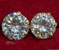 Close-up image of the pair of round, clear cubic zirconia stud earrings set in yellow gold prong settings on a red background.