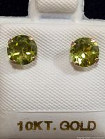 Pair of round faceted light green peridot stud earrings set in 10K gold prong settings displayed on white textured card.
