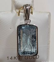 Close-up image of a rectangular faceted aquamarine set in 14K gold pendant with bail. Backdrop shows '14KT GOLD' marking.