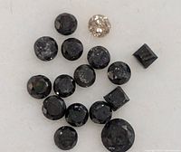 Close-up of a cluster of small faceted black diamonds and one light-colored diamond on a white surface, showing various cuts.