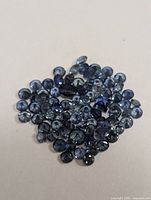 Photo of numerous small loose round-cut blue sapphires on a white background showing their varied blue hues and reflective properties.