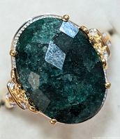 Close-up of oval dark green emerald set in silver and gold-tone ring showing gemstone facets and setting details.