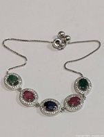 Top view of the silver bracelet showing the oval green emeralds, red rubies, and the blue sapphire gemstones, each surrounded by small clear stones.