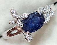 Close-up of silver rhodium plated ring with an oval blue sapphire center stone and butterfly-shaped accents encrusted with small clear stones.