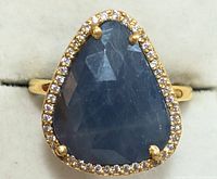 Close-up image of silver ring with large triangular blue sapphire center stone surrounded by small clear stones set in yellow gold band.