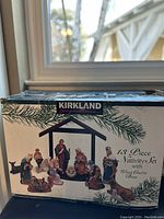Box of Kirkland Signature 13 Piece Nativity Set with Wood Creche Base showing the assembled figurines in the nativity scene including animals and human figures, displayed on wooden creche base.