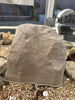 Tan colored faux stone boulder on a bed of small gravel with other rocks in background.