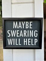 Photo of a black-framed rectangular novelty sign with white text 'MAYBE SWEARING WILL HELP'.