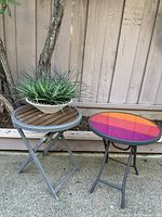 Photo of two small round patio tables, one with a wooden slatted top and the other with a colorful striped top, next to a tree and fence outdoors