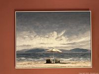 Full view of framed print showing beach chairs and umbrella on shore with ocean and mountains and cloudy sky.