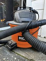 Close-up photo of an orange and black Rigid shop vac showing flexible black hose attached and power cord wrapped on top.